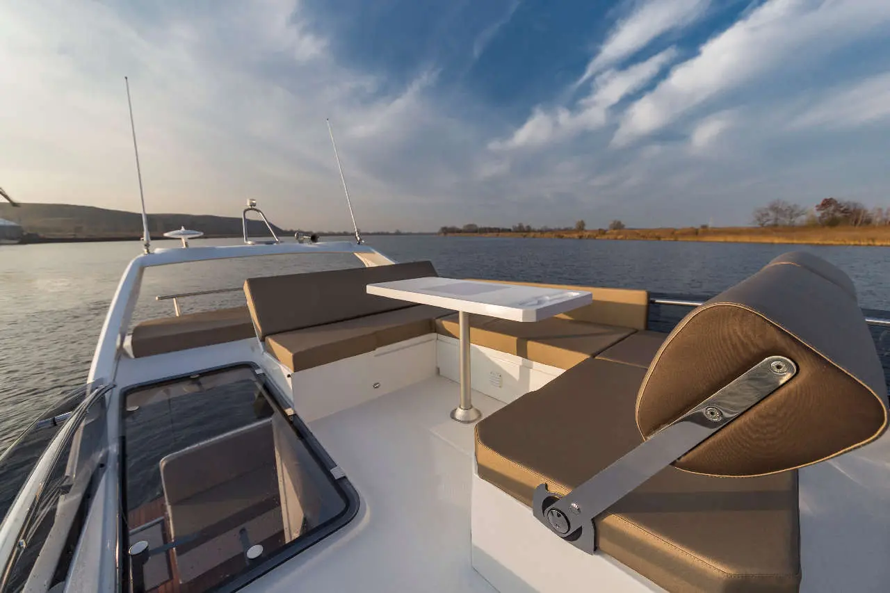 A spacious boat deck featuring cushioned seating, a small table, and scenic views of a river under a clear sky.