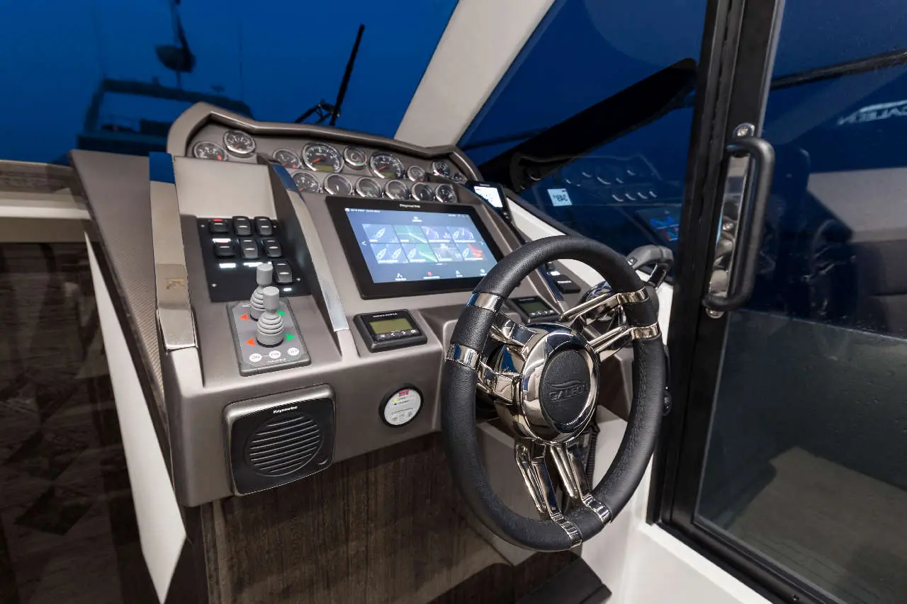 Boat cockpit featuring a steering wheel, digital display, controls, and gauges, with a window showing a dark, cloudy sky.