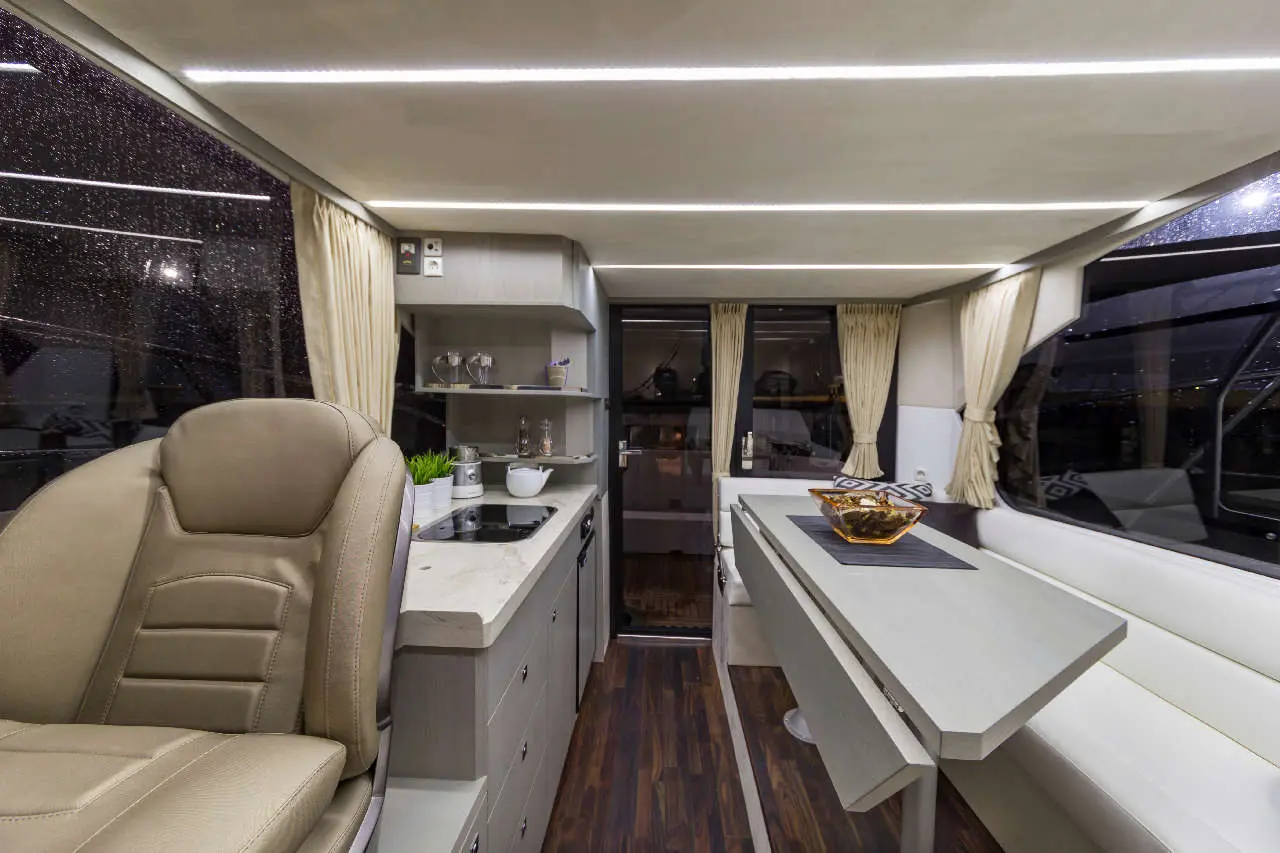 Modern boat interior with a beige captain's chair, kitchenette, and dining table under illuminated ceiling, featuring large windows.