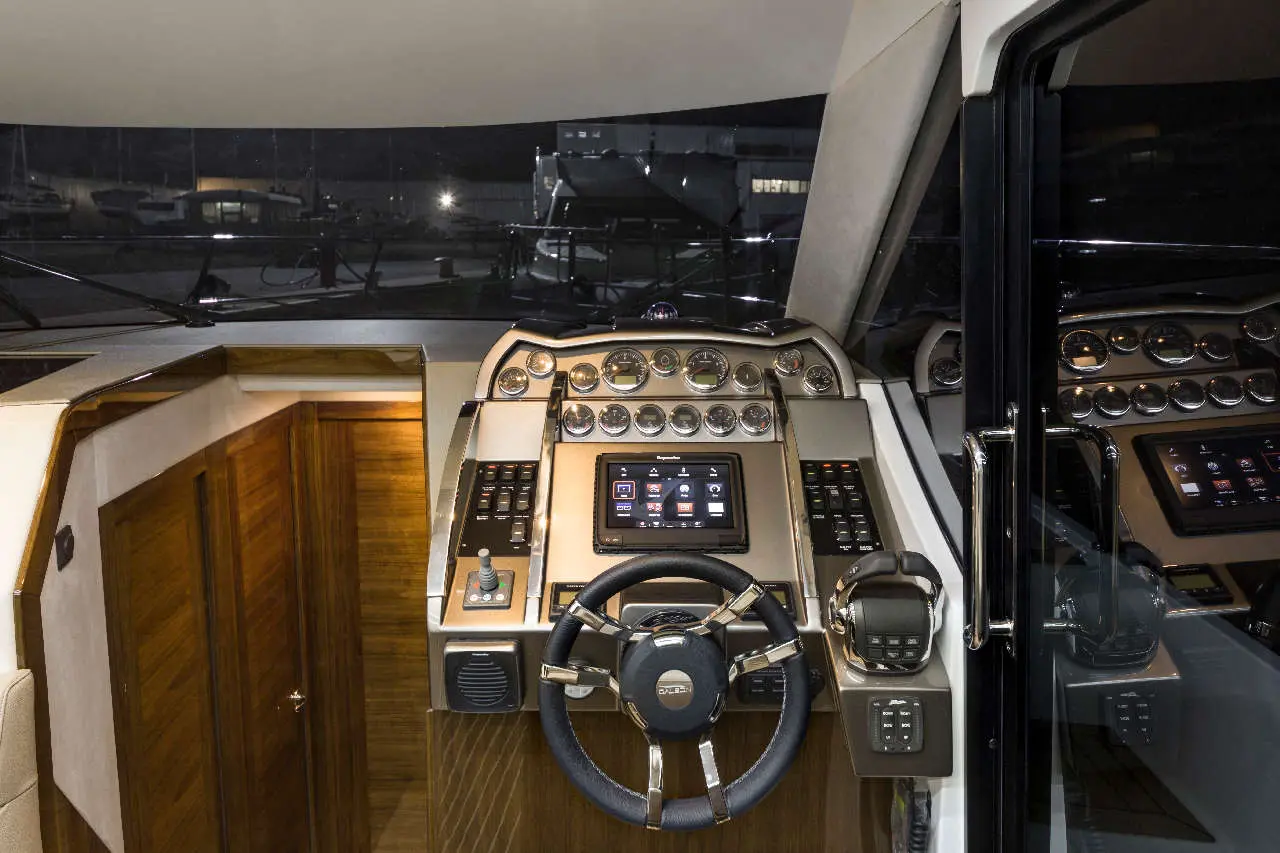 Luxury yacht helm with a sleek steering wheel, digital displays, dials, and wooden interior, illuminated in evening light.