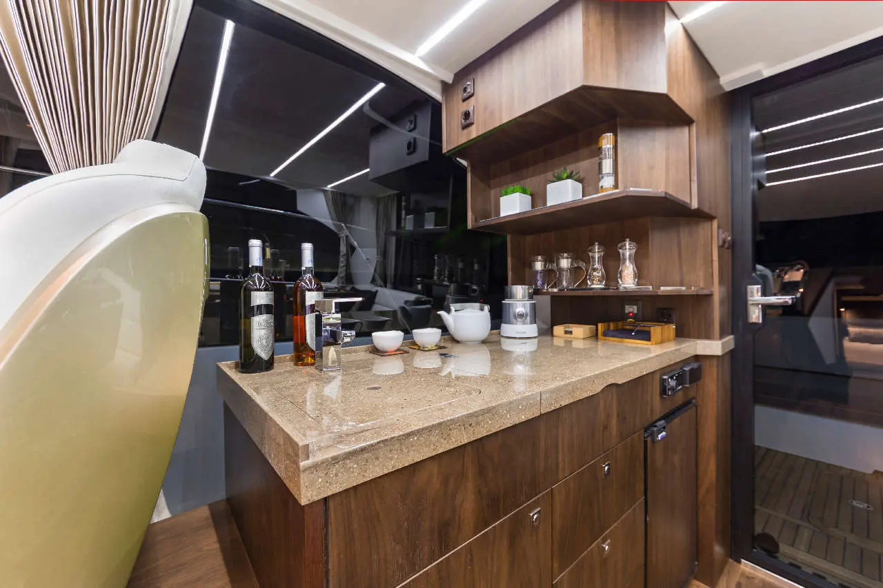 Modern yacht galley with a stone countertop, wine bottles, tea set, kitchen appliances, and sleek wooden cabinetry.