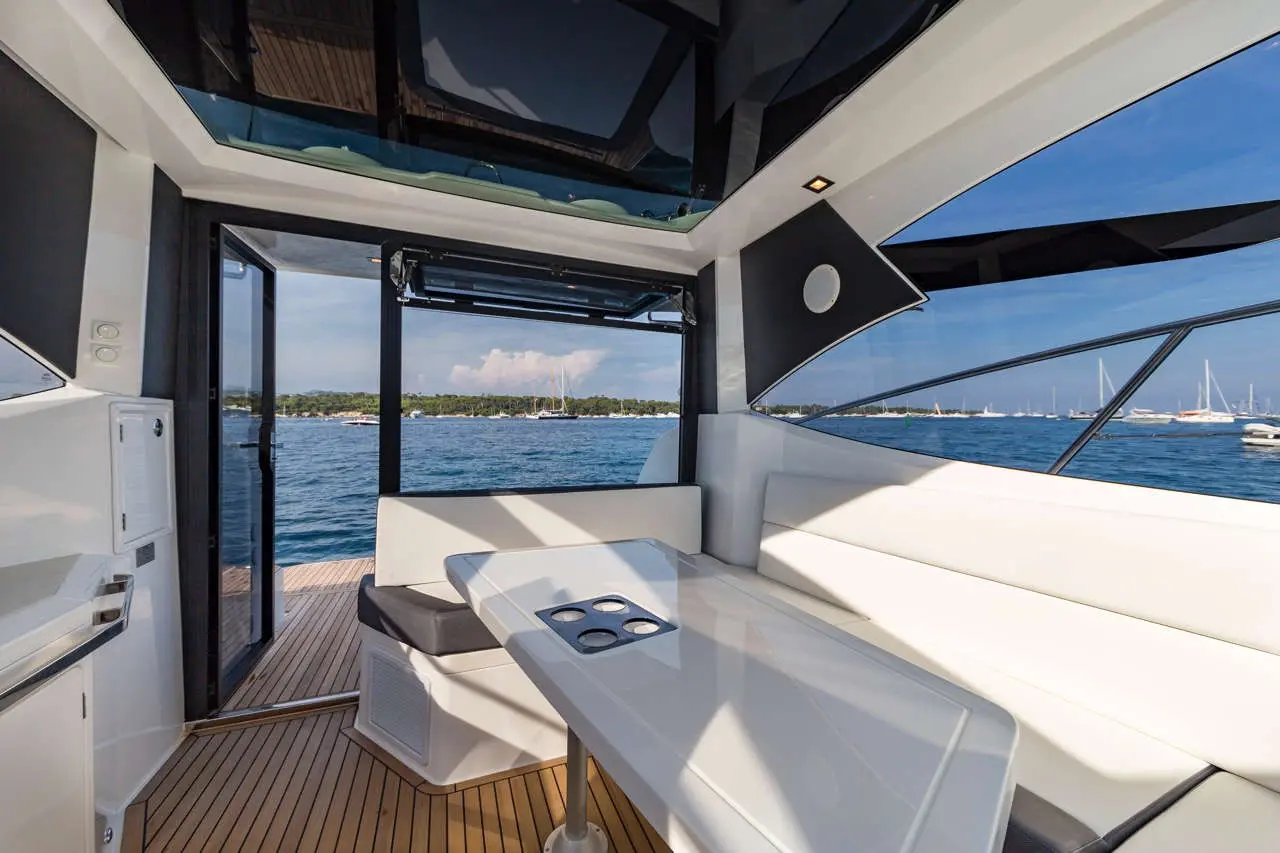 Interior view of a modern boat with large windows, a dining table, and a scenic waterfront in the background.