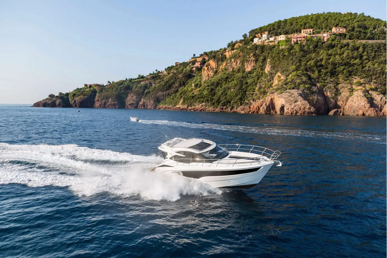 A white motorboat speeds through clear blue waters near a green hillside with a coastal village in the background.