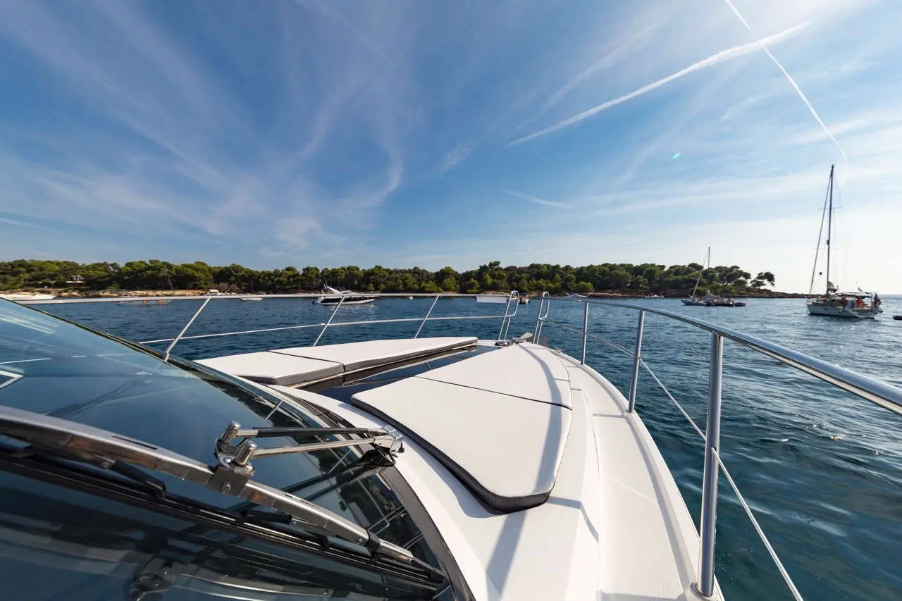 A motorboat's bow view reveals calm waters, nearby yachts, and a scenic green shoreline under a blue sky.