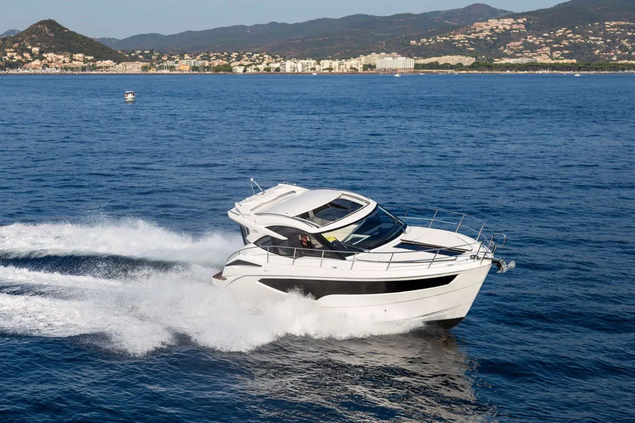 A sleek white motorboat speeds across calm blue waters with a coastal city and mountains in the background.