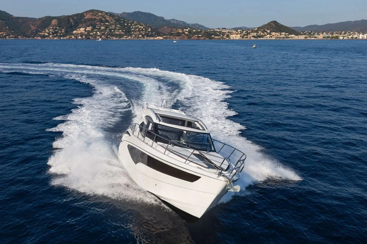 A sleek white motorboat creates a wake as it speeds through calm blue waters, with mountains and a coastline in the background.