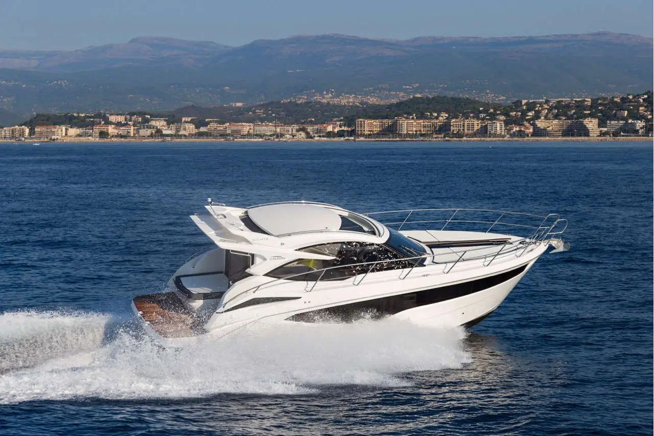 A sleek white yacht speeds through blue waters, with mountains and a coastal town visible in the background.