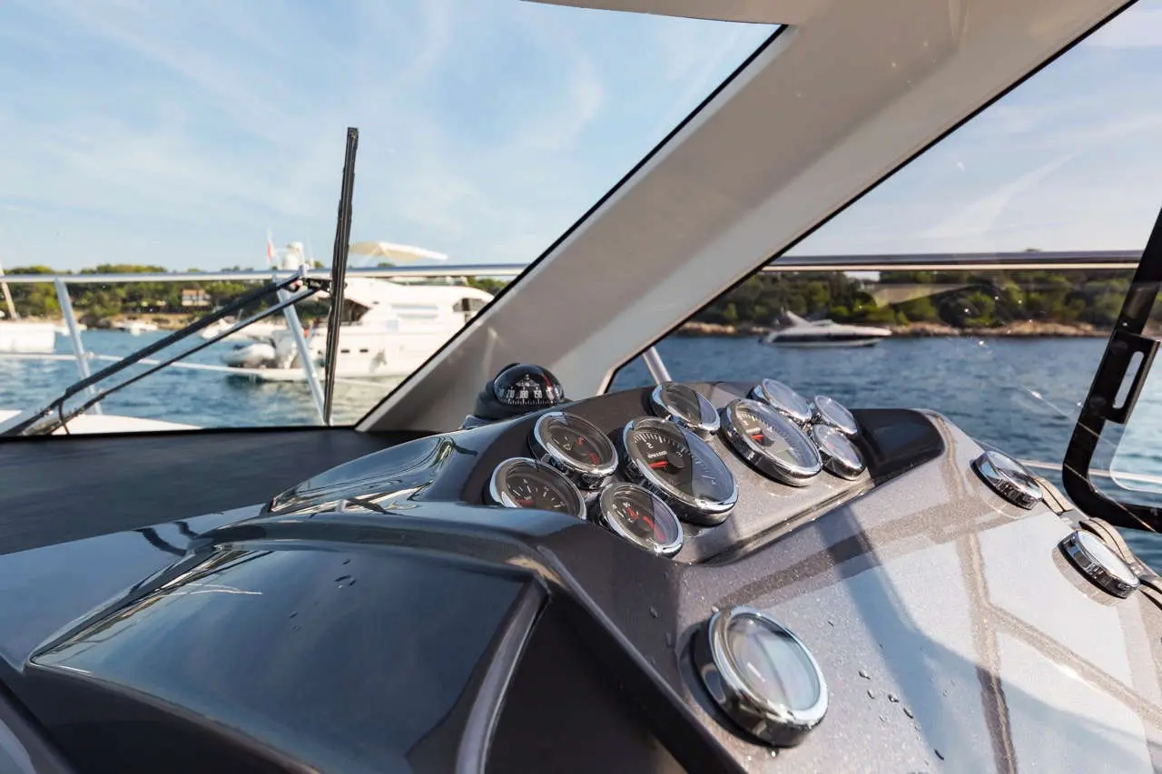 View of a yacht's dashboard with chrome gauges, a compass, and boats on a calm water backdrop under a blue sky.