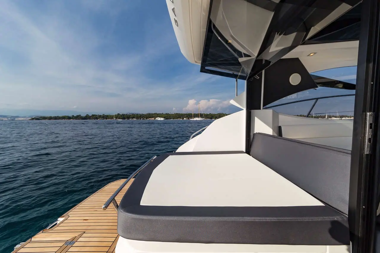 A view of a yacht's sun deck with plush seating, overlooking calm waters and a distant coastline under a blue sky.