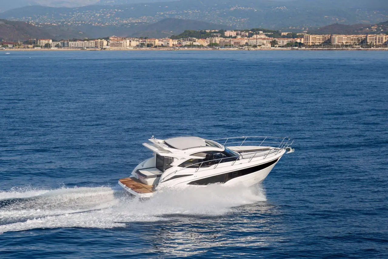 A sleek white motorboat accelerates through calm blue waters with a coastal city and mountains in the background.