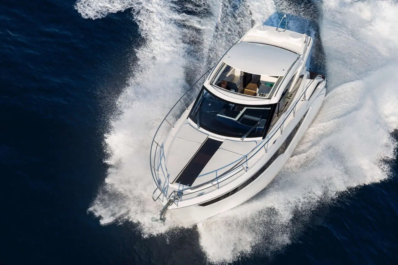 A white luxury motor yacht glides through deep blue water, creating waves while a person is visible at the helm.