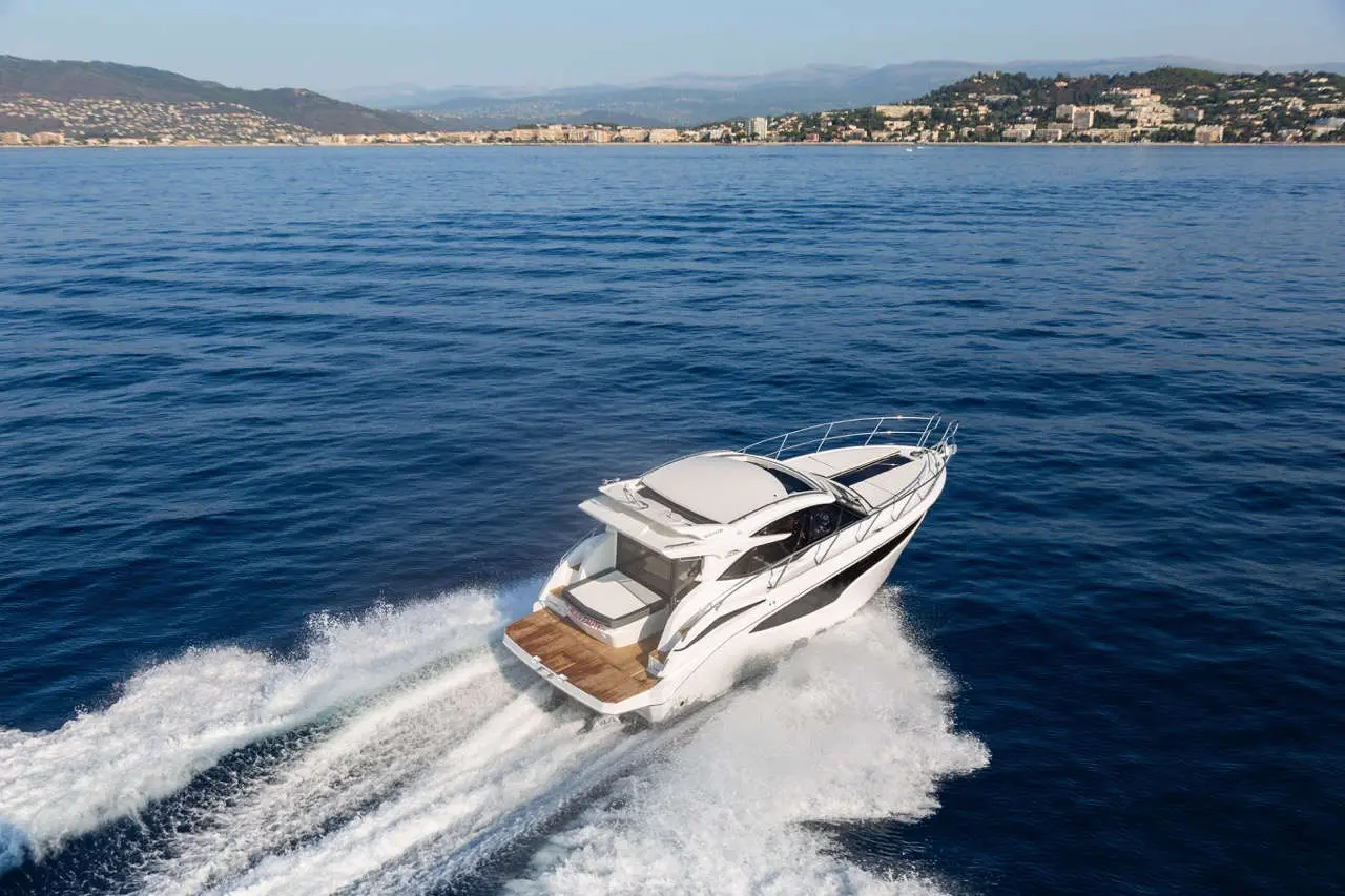 A sleek white motorboat speeds across blue waters, with a coastal town visible in the background under clear skies.
