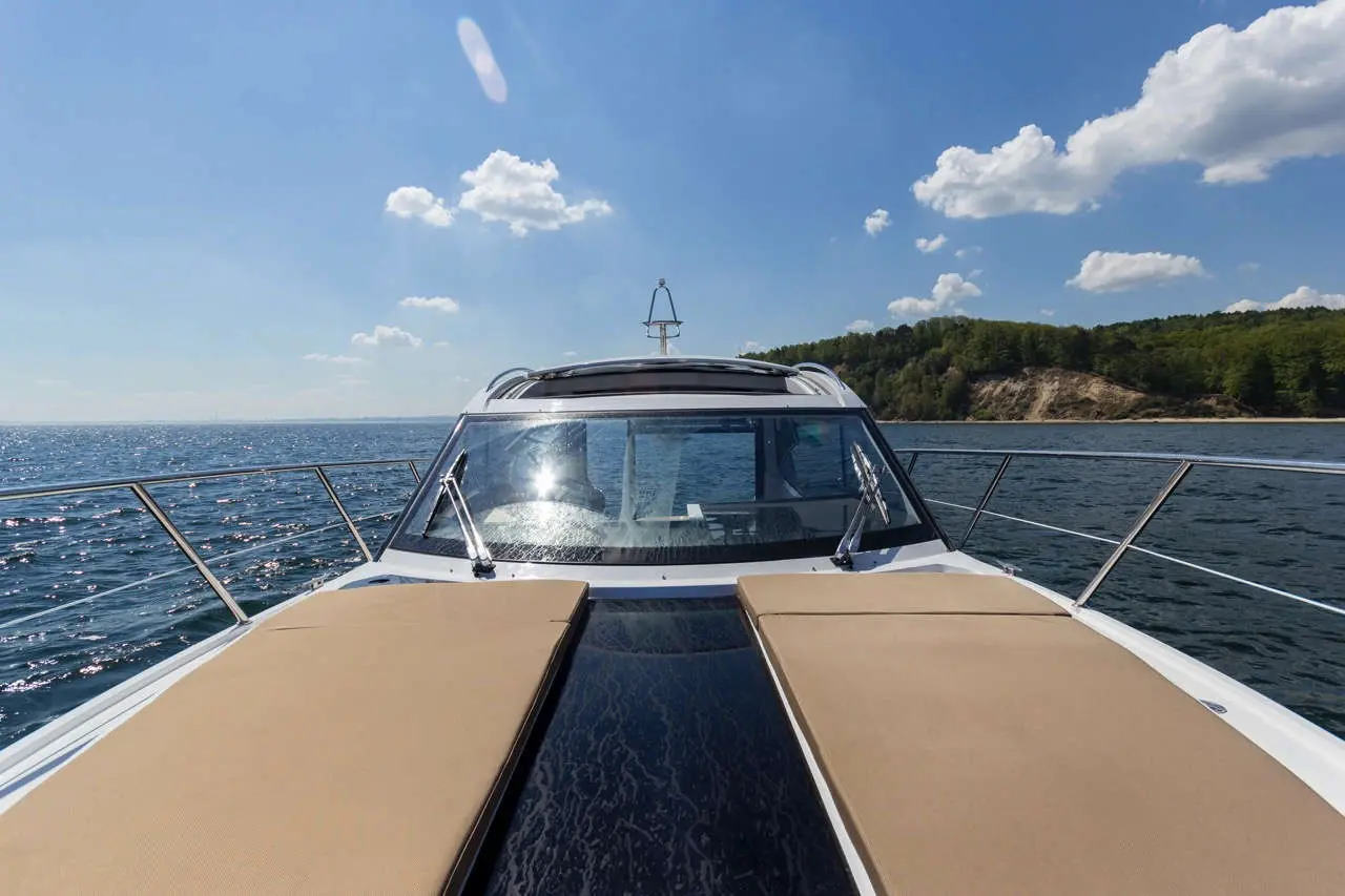 View from the bow of a boat, showcasing the ocean, clouds, and a forested shoreline under bright sunlight.