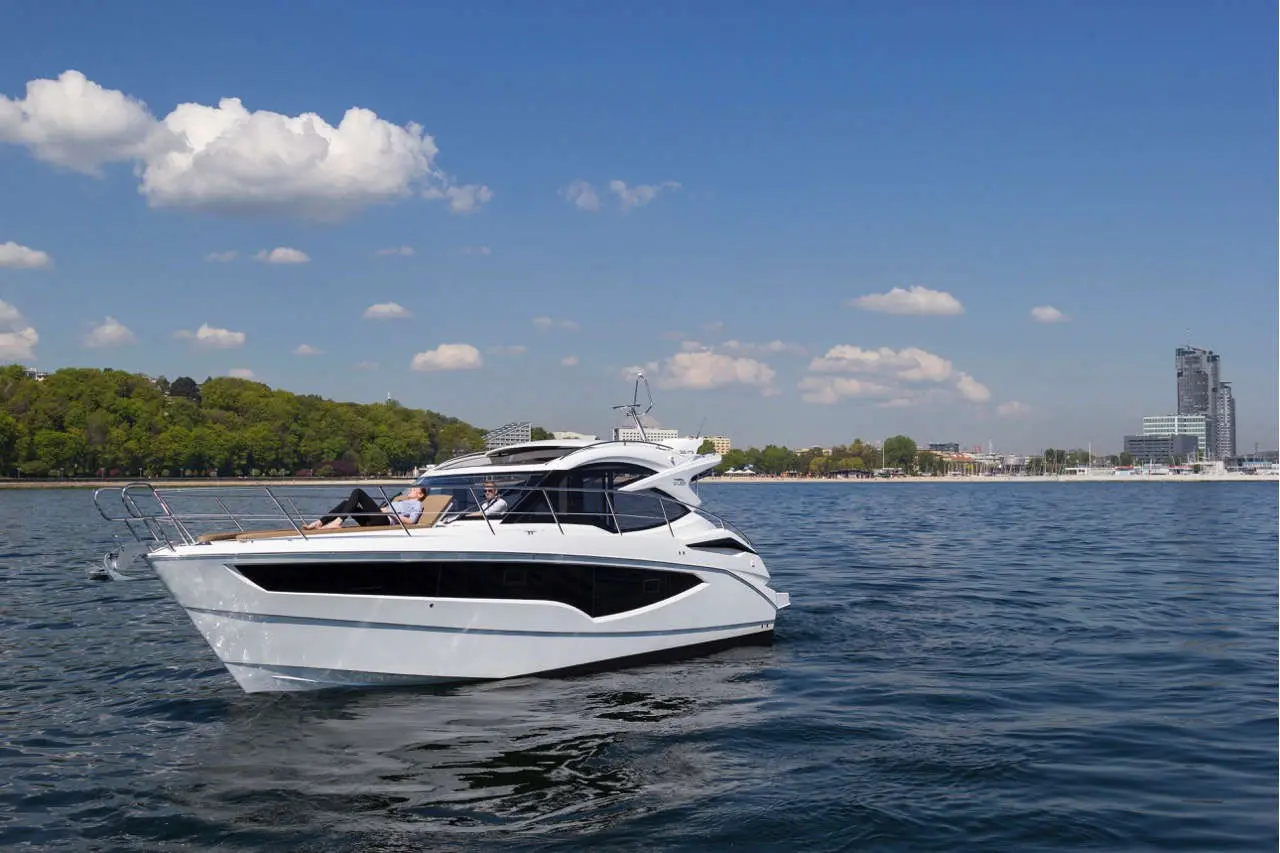 Luxury motor yacht cruising on calm water, with two people relaxing on board, city skyline and lush greenery in the background.