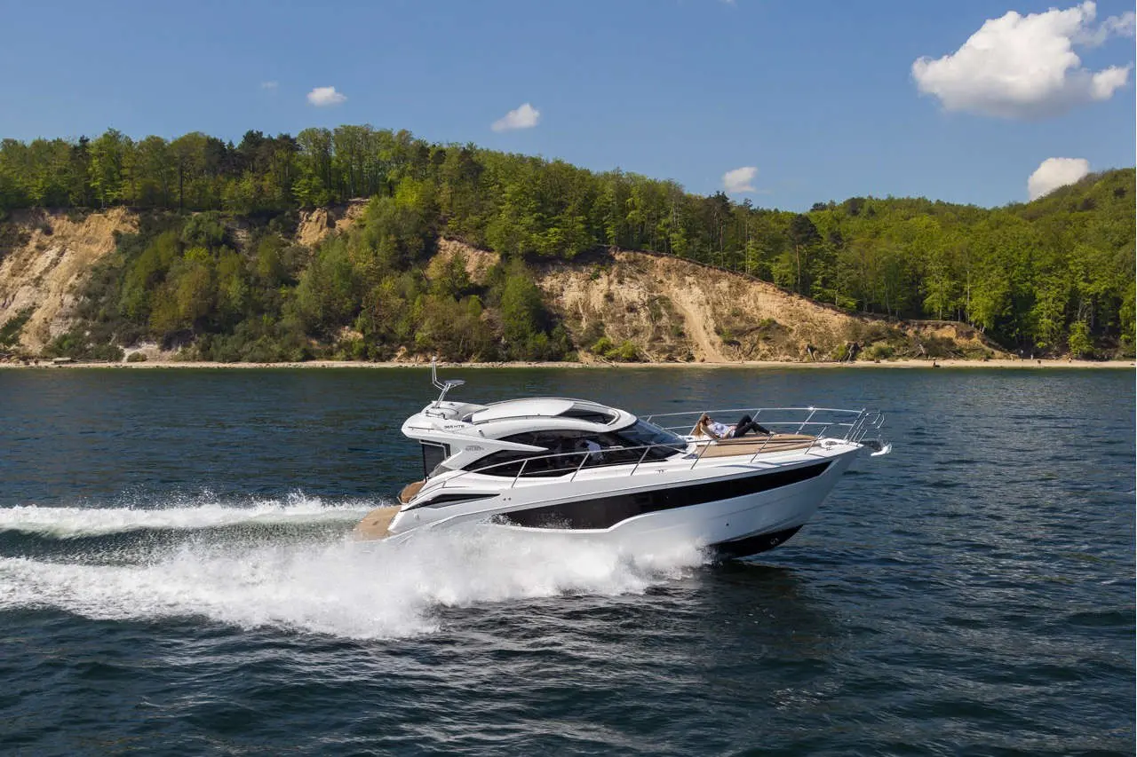 Luxury boat cruising on calm waters near a lush green hillside, with two people relaxing onboard under a clear blue sky.