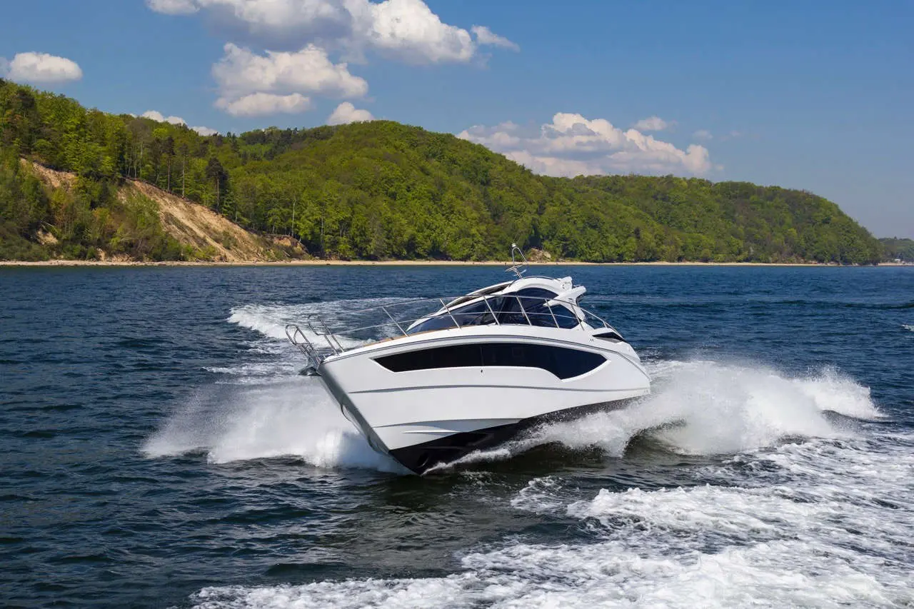 A sleek white motorboat cuts through blue waters, creating splashes near a lush green shoreline under a clear sky.