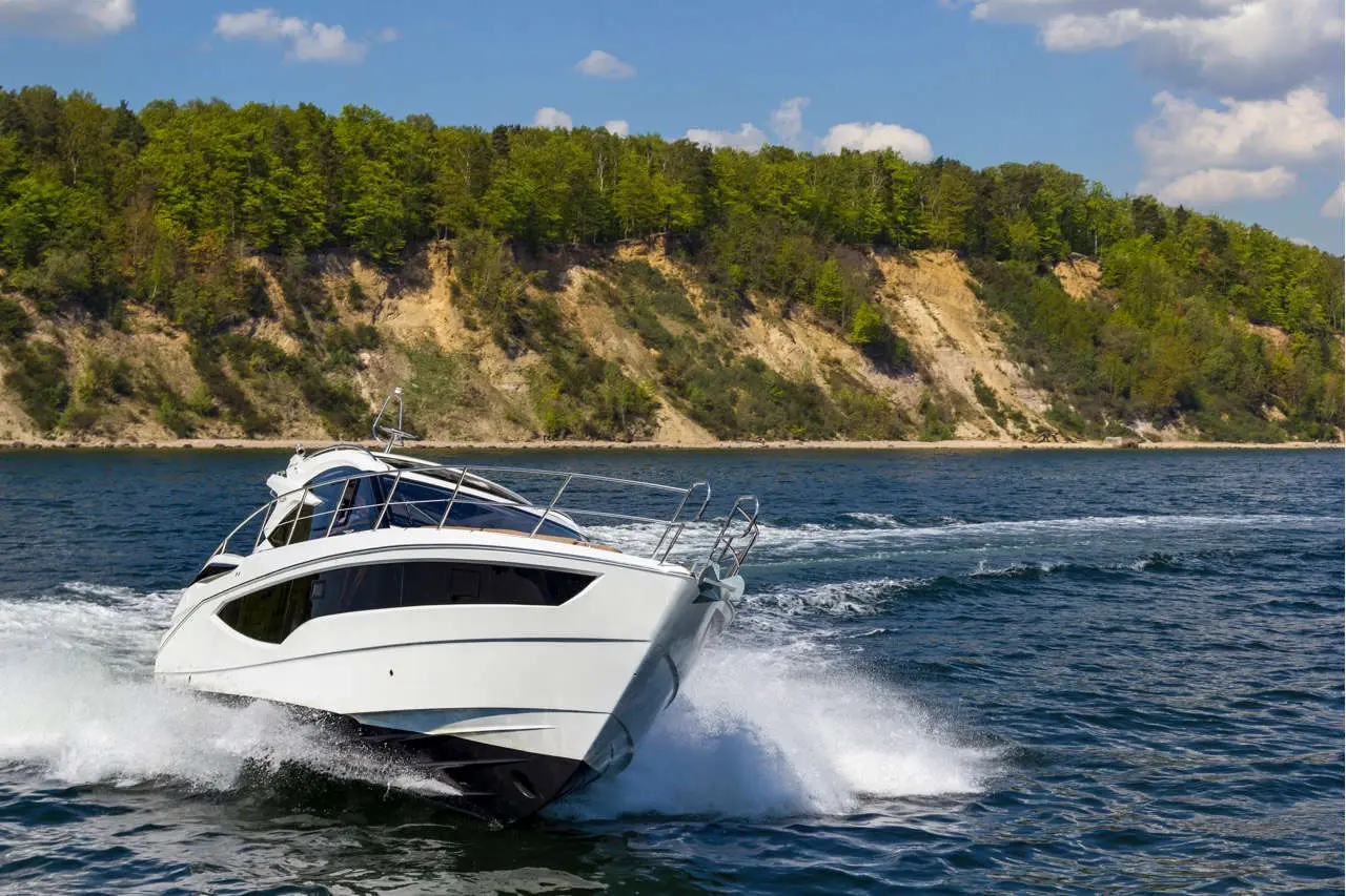A sleek white motorboat speeds across blue water, leaving a wake near a green forested cliff under a bright sky.