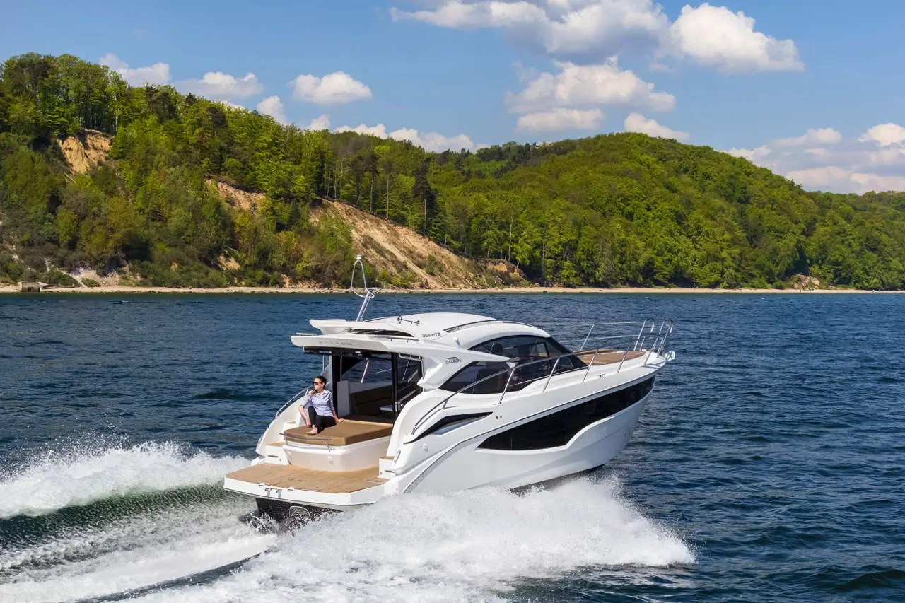 A sleek white motorboat cruises through clear blue water with a person lounging on the deck against a backdrop of lush green hills.