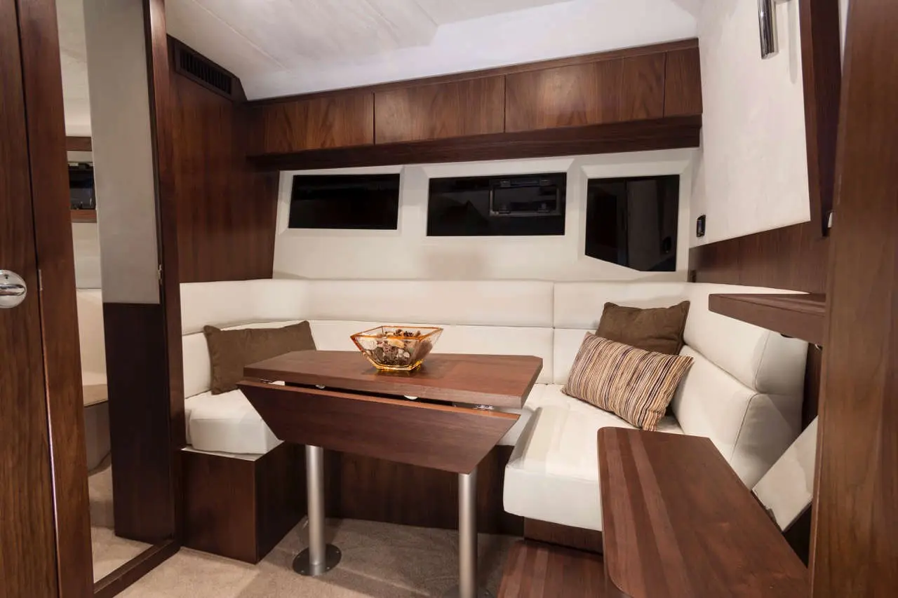Modern boat interior featuring a wooden table, white seating, a bowl of decorative items, and large windows for natural light.
