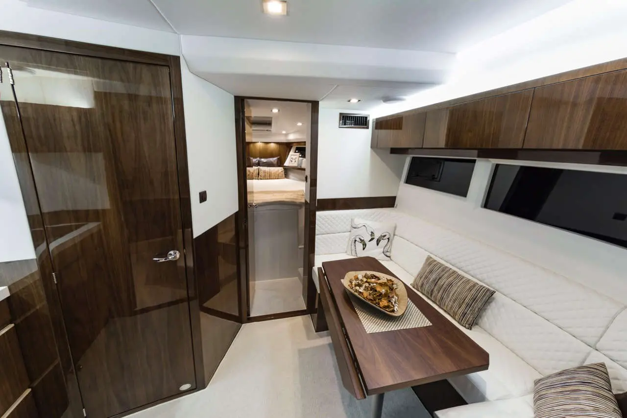 Luxury yacht interior with a dining area, white leather seating, wood accents, and a doorway leading to a bedroom.