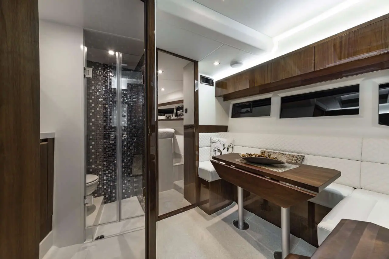 Modern boat interior featuring a dining area with white seating, wooden table, and a bathroom with glass door and tiled shower.
