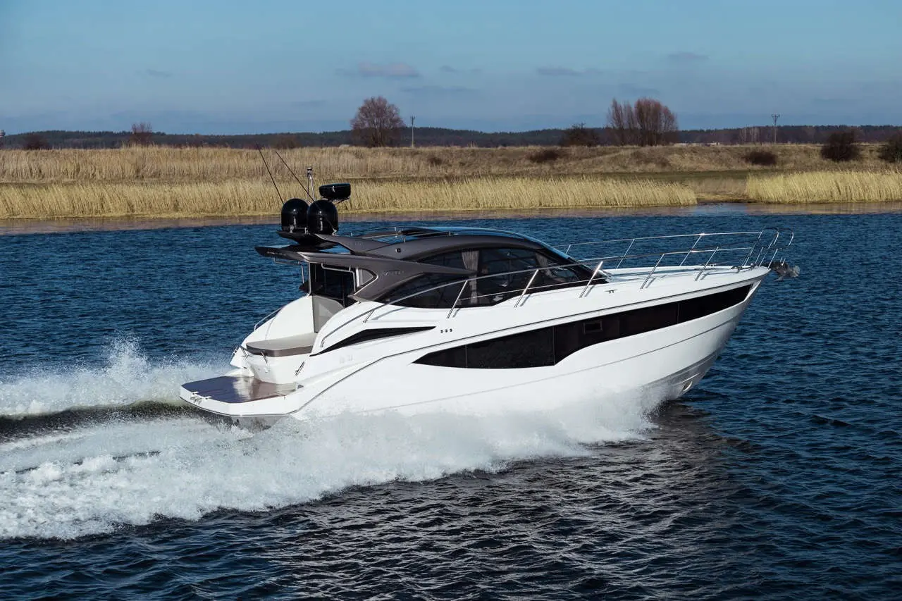 A sleek white motor yacht glides over water, creating waves, with green reeds visible along the shoreline under a clear blue sky.