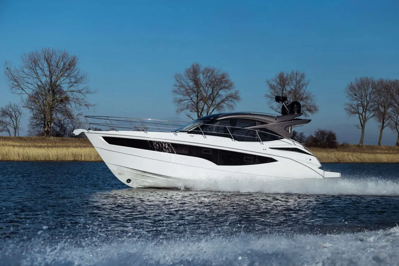 A sleek white motor yacht glides across calm water, with trees in the background under a clear blue sky.