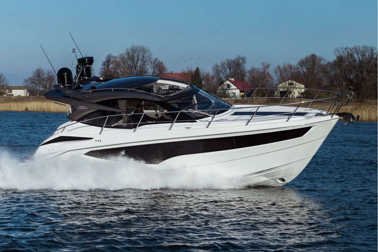 A sleek white motorboat glides through water, creating waves, with trees and houses visible in the background.