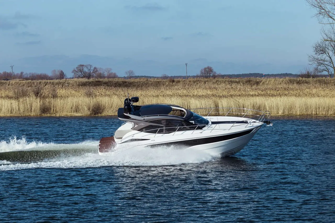 A sleek white yacht speeds through calm water, leaving a wake, with tall grass and trees in the background under a clear sky.