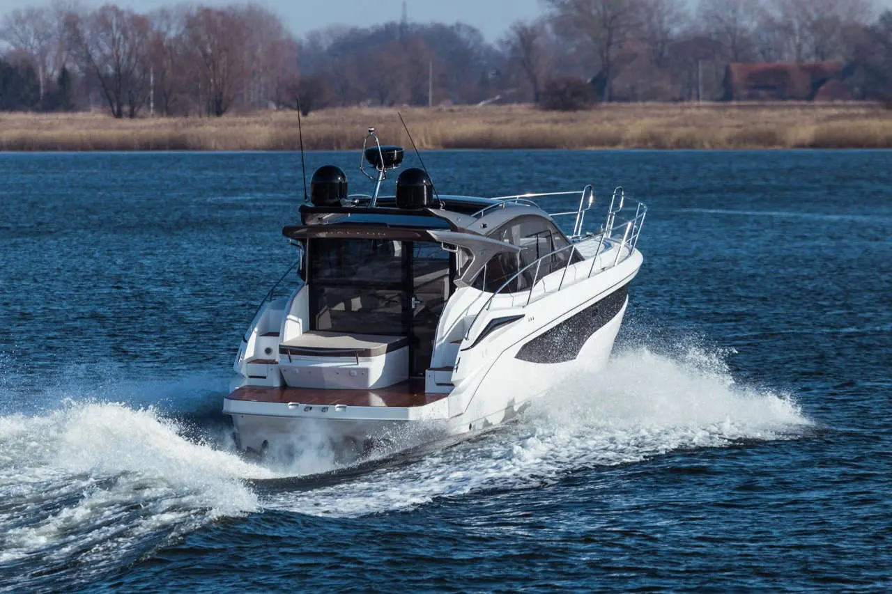 A sleek white motorboat speeds through blue water, creating splashes, with trees visible on the shore.