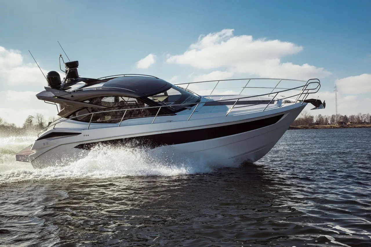 A sleek white motorboat glides over calm water, creating splashes, with blue skies and soft clouds in the background.