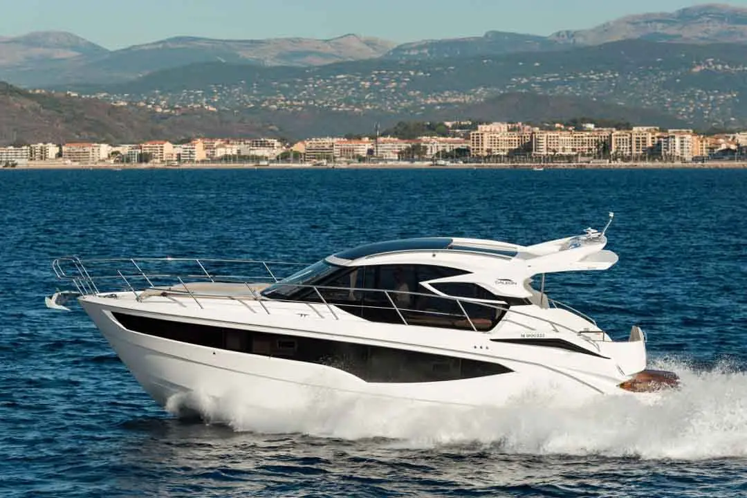 A sleek white motor yacht glides through blue waters, with a coastal city and mountains in the background.