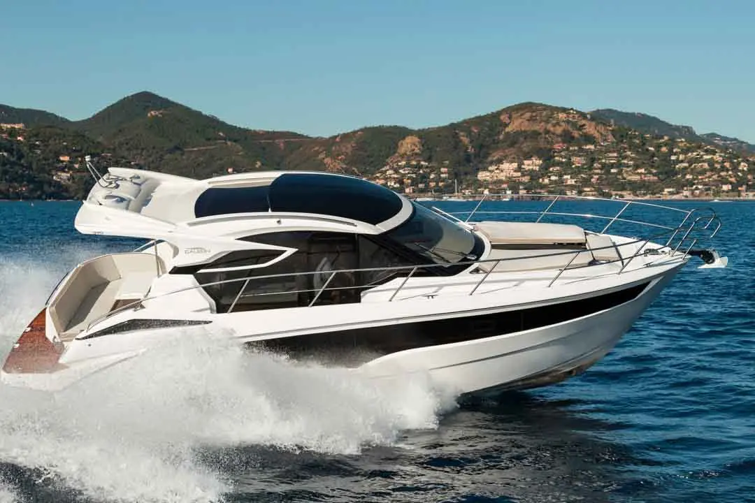A sleek white motorboat glides through clear blue water, with green mountains and a coastal village in the background.
