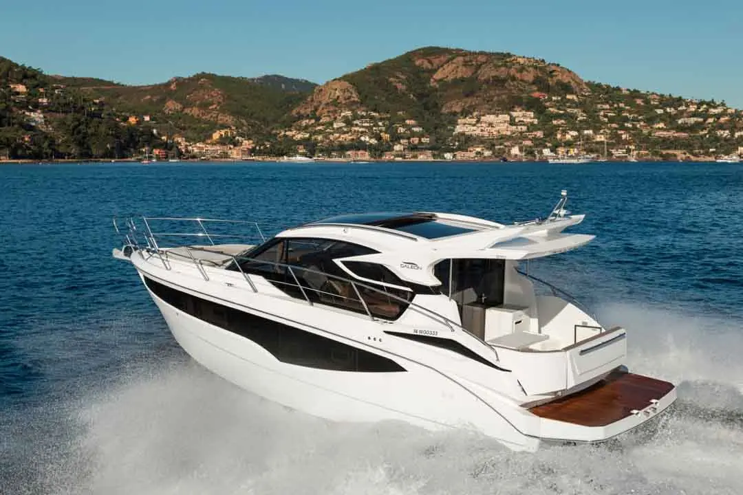 A sleek white and black motorboat speeds through blue water, with a scenic hillside and homes in the background.