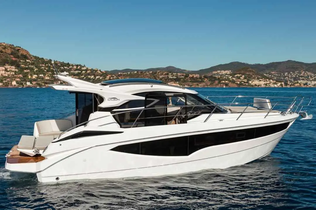 A sleek white yacht with cabin windows and a blue sky backdrop, cruising through calm waters with distant hills.