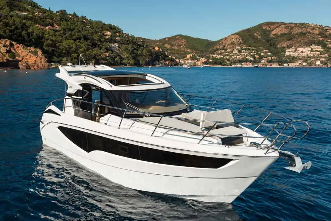 A modern white motorboat is cruising in clear blue water, with lush green hills and buildings in the background.