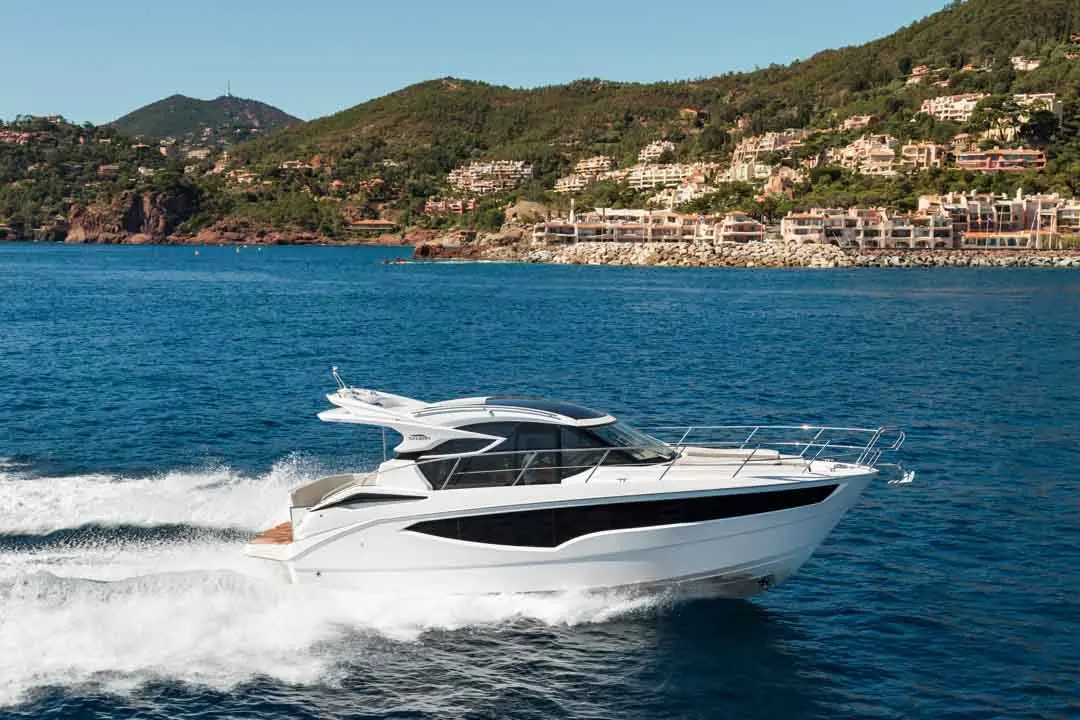 A sleek white motor yacht gliding through blue waters, scenic hills and luxury homes visible in the background.