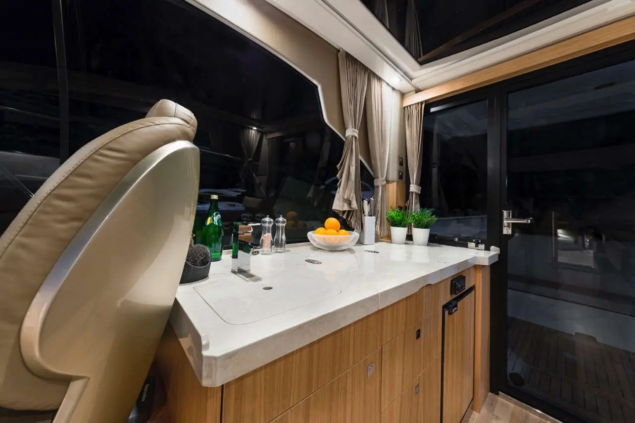 Modern yacht interior featuring a sleek kitchen area, fresh fruit bowl, glassware, and a leather chair overlooking the water.
