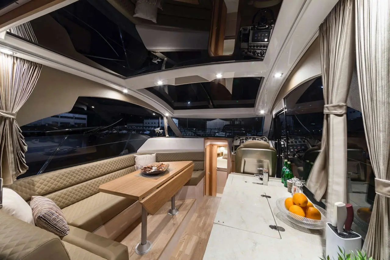 Modern yacht interior featuring a dining area, kitchenette, and large windows. An arrangement of oranges is on the counter.