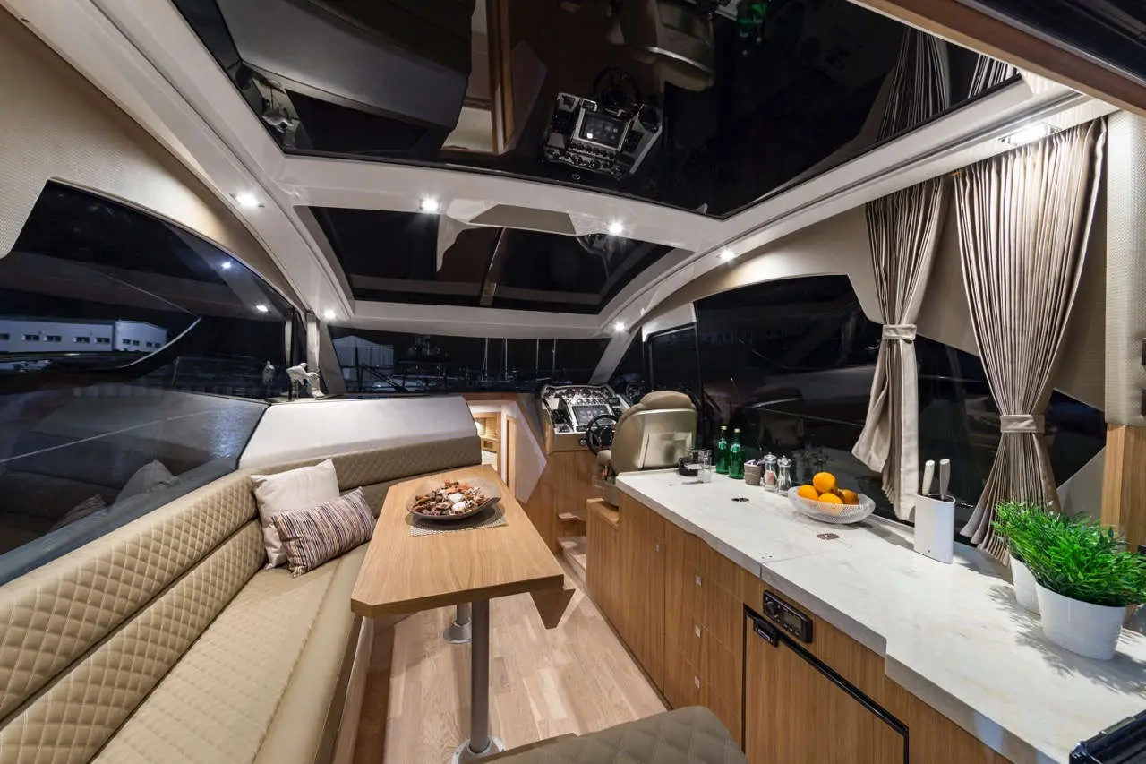 Modern yacht interior featuring a cozy seating area, sleek kitchen counter with fruit, and navigation control station, illuminated at night.