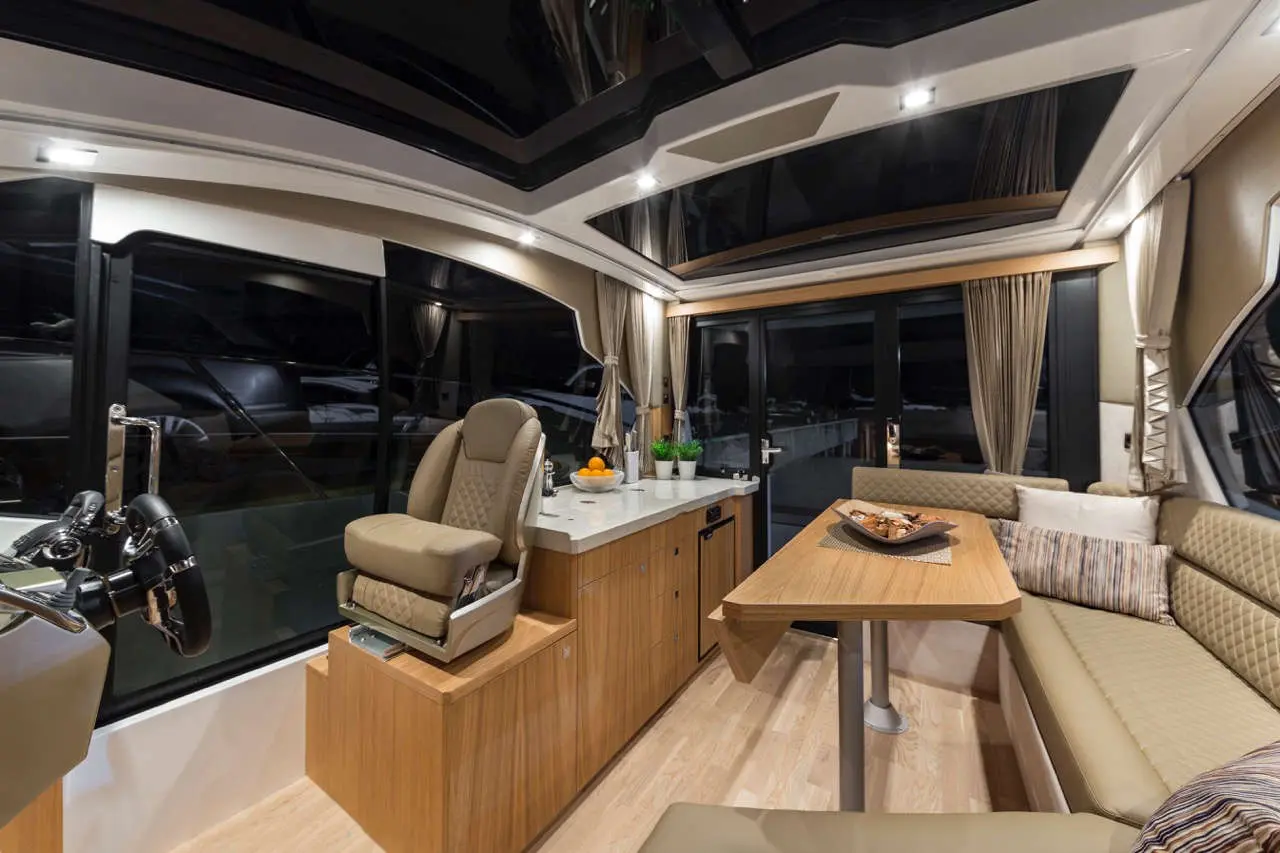 Interior of a luxury boat showing a seating area, helm chair, dining table, large windows, and decorative fruit bowl.