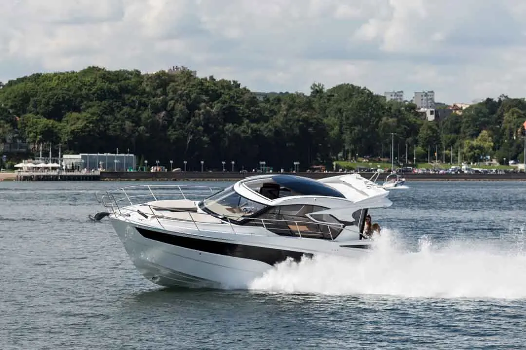 A luxurious motorboat speeds through water, creating a spray, with a lush green landscape and buildings visible in the background.