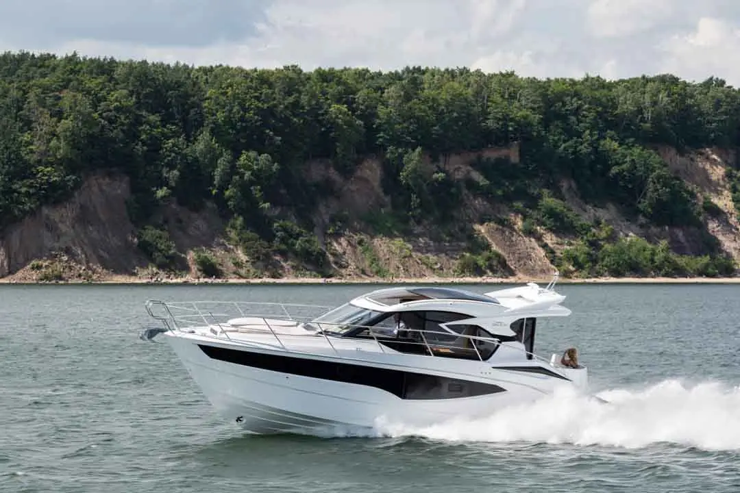 A sleek white motorboat speeds across calm waters, with a lush green hillside in the background and a clear sky overhead.