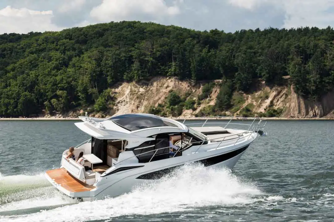 Luxury motorboat cruising through calm waters near a forested shoreline, with two people enjoying the ride.