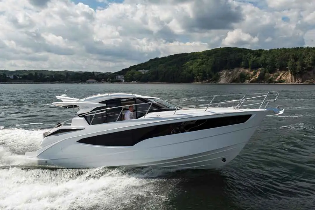 A sleek white motorboat glides through calm waters, with a person at the helm and green hills in the background.
