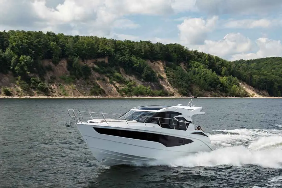 A white motor yacht navigates through calm waters, surrounded by lush green hills under a partly cloudy sky.