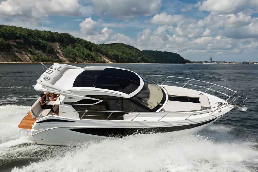 A sleek white motorboat glides through water, with a woman seated on the deck against a backdrop of green hills and blue skies.