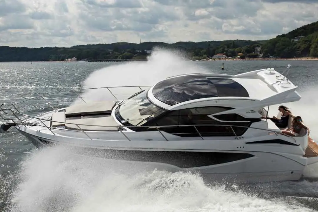 A sleek white motorboat speeds over water, with two women enjoying the ride on the back. Lush greenery is visible in the background.