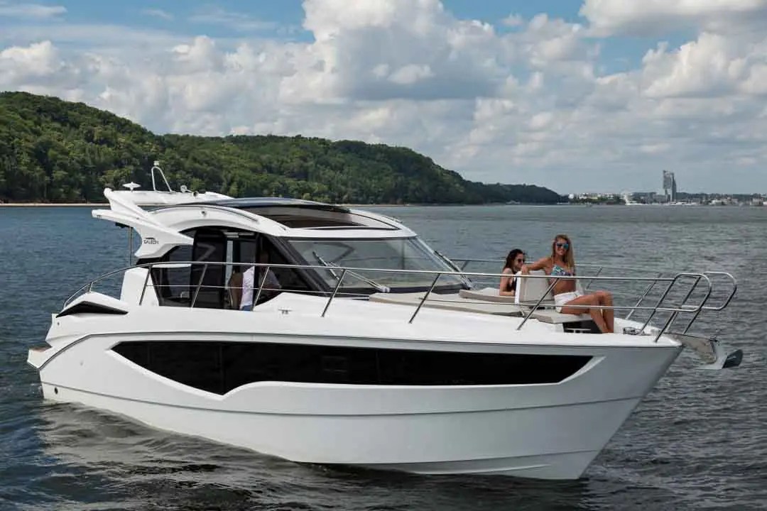A white luxury yacht on water with two women enjoying the sun, surrounded by green hills and a bright blue sky.