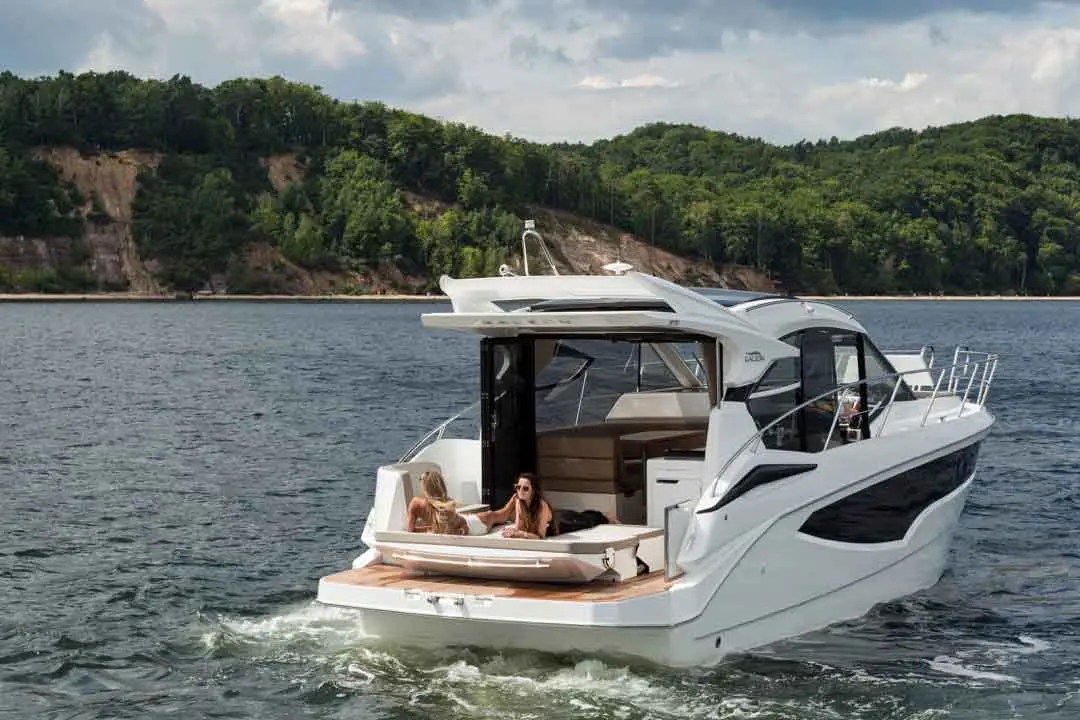 Two women relax on a luxury motorboat in a lake, surrounded by lush green hills and a cloudy sky.